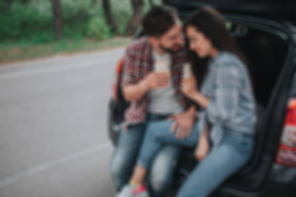 Blurred picture of young couple sitting in trunk and holding rolls in their hands. Guy is looking at girl. She is leaning to his shoulder. They look happy. - Фото, изображение