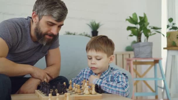 Serious preschool child is playing chess with his parent thinking about next move and moving chesspieces while his father is teaching him game tactics. - Filmati, video