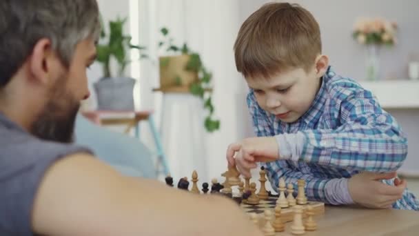 Clever little boy is learning to play chess moving chesspieces on board and enjoying playing with his dad. His fathers bearded face is in foreground. - 映像、動画