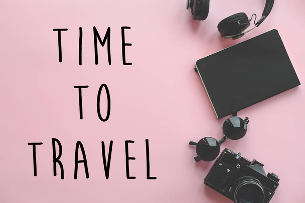 Χρόνος ταξιδιού κείμενο σε κομψό μαύρο φωτογραφική, ακουστικά, γυαλιά ηλίου και σημειωματάριο σε μοντέρνα ροζ φόντο, επίπεδη lay. μοντέρνο κουρασμένος χίπι ταξίδια και περιπλάνησης εικόνας. - Φωτογραφία, εικόνα