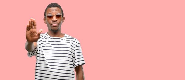Uomo nero africano con gli occhiali da sole infastidito dal cattivo atteggiamento facendo segno di stop con la mano, dicendo di no, esprimendo sicurezza, difesa o restrizione, forse spingendo
 - Foto, immagini