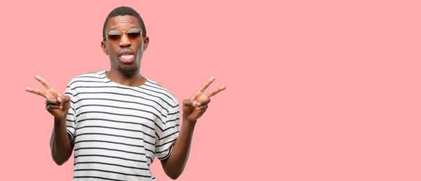 Afrikkalainen musta mies yllään aurinkolasit katselee kameraa osoittaa tong ja tehdä voitto merkki sormilla
 - Valokuva, kuva
