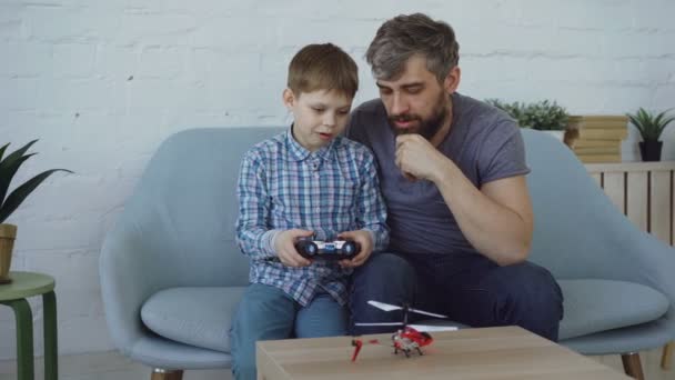 Cute child is holding transmitter and controlling flying helicopter while his father is trying to catch it with his hands. Family members are laughing and having fun. - Video