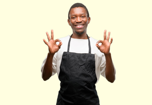Afrikai férfi üzlet tulajdonosa viselt kötény csinál ok jel gesztus, mindkét kezével, kifejező meditáció és relaxáció - Fotó, kép