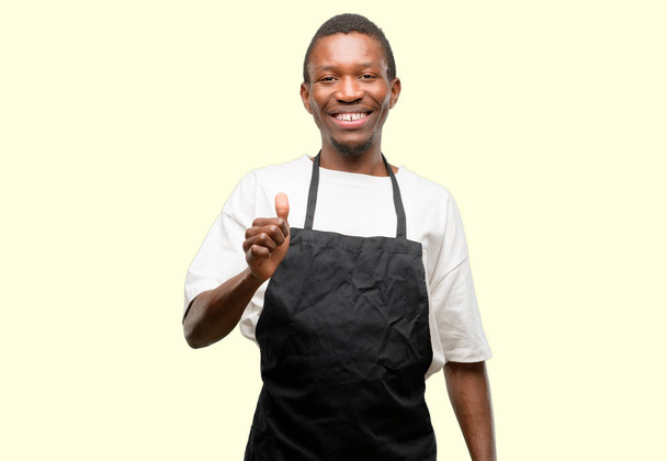 Africano proprietário loja homem vestindo avental sorrindo amplamente mostrando polegares até gesto para câmera, expressão de gosto e aprovação
 - Foto, Imagem