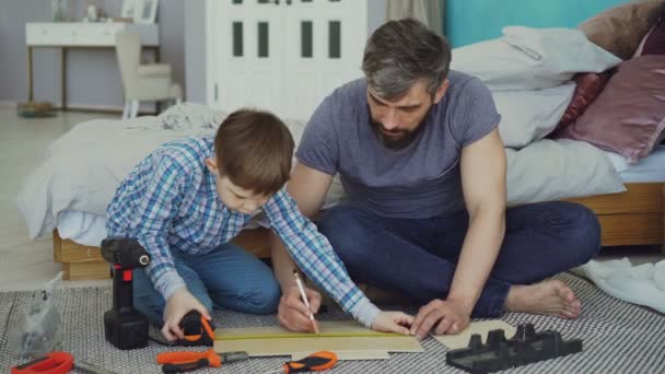Father and son are measuring piece of wood with measure-reel getting ready to construct something together inside house. Different tools and modern furniture are visible. - Filmmaterial, Video