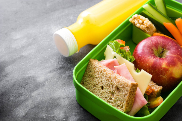 Здоровая школьная коробка для обеда: Сэндвич, овощи, фрукты и сок на черном камне
 - Фото, изображение
