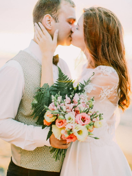 The groom is holding the wedding bouquet of the roses while kissing with the beautiful bride. - Photo, image