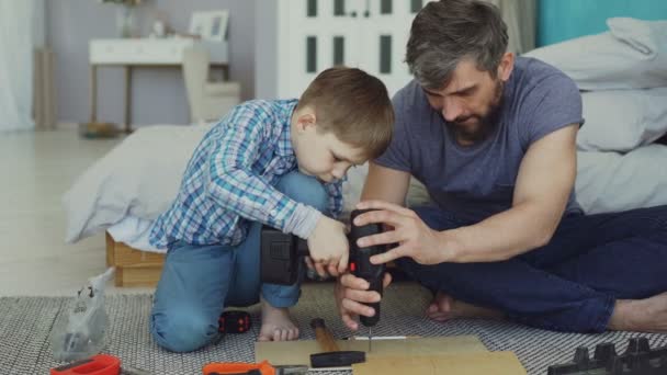 Careful father is teaching his son to work with electric screwdriver while son is trying to use screw gun and fix screw in pieces of wood. Construction and family concept. - Кадри, відео