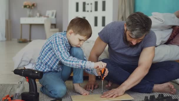 Serious small boy is concentrated on putting screw in pieces of wood with screwdriver while his father is helping him holding wooden sheet. Childhood and construction concept. - Кадры, видео
