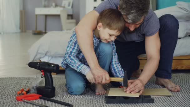 Father bearded man is teaching his son how to use hammer driving nail in piece of wood together sitting on floor at home. Instruments, tools and furniture are visible. - Кадри, відео