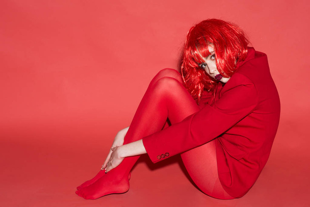 Fashion concept. Lady looking at camera and sits on floor. Woman with makeup and red wig posing in total red outfit. Girl on mysterious face in red formal jacket and tights, red background. - Fotó, kép