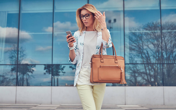 Fashionable female blogger in stylish clothes and glasses with a handbag, holds a smartphone, standing against a skyscraper. - Photo, Image