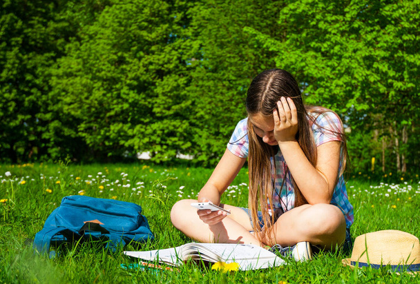 Студент учится в парке со смартфоном. Радостная счастливая студентка-подросток сидит и читает снаружи университетского городка или парка
. - Фото, изображение
