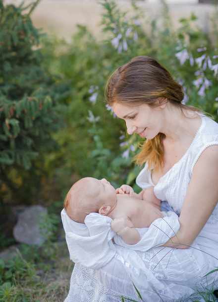 Μια όμορφη νεαρή μητέρα με ένα μωρό στην αγκαλιά της στέκεται στον κήπο. - Φωτογραφία, εικόνα