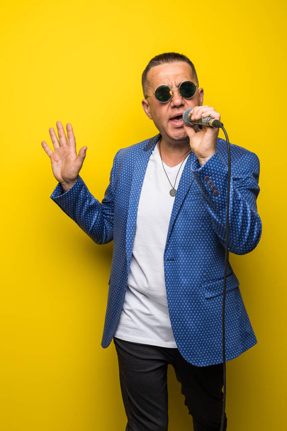 Porträt eines reifen Mannes mittleren Alters im Anzug, der isoliert auf gelbem Hintergrund über das Mikrofon singt. Sänger-Konzept. - Foto, Bild