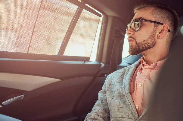 Успешный стильный молодой бизнесмен в сером костюме и розовой рубашке, катающийся на заднем сидении в роскошном автомобиле
. - Фото, изображение