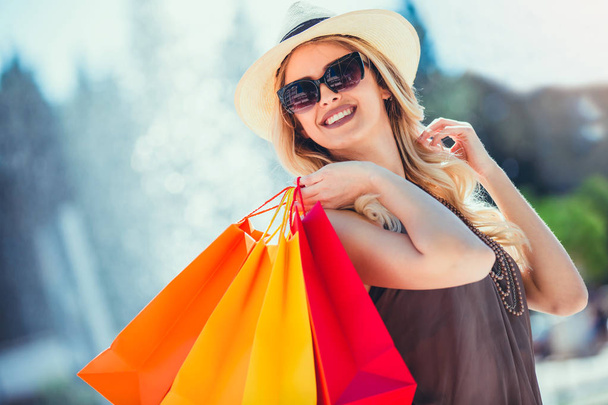 Blondi nainen aurinkolasit tilalla ostoskassit ja hymyillen kävellessään
 - Valokuva, kuva