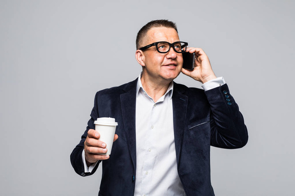 Senior-Geschäftsmann hält Kaffeetasse, während er auf dem Handy vor weißem Hintergrund spricht - Foto, Bild