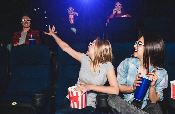 Funny picture of girls sitting together in cinema hall. They are looking at the boy that sits behind them. Blonde girl is trying to reach guy with her hand. Young woman are laughing. - Photo, Image