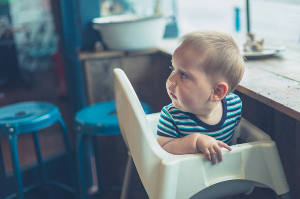 A little baby is sitting in a high chair by the window in a cafe - Foto, imagen