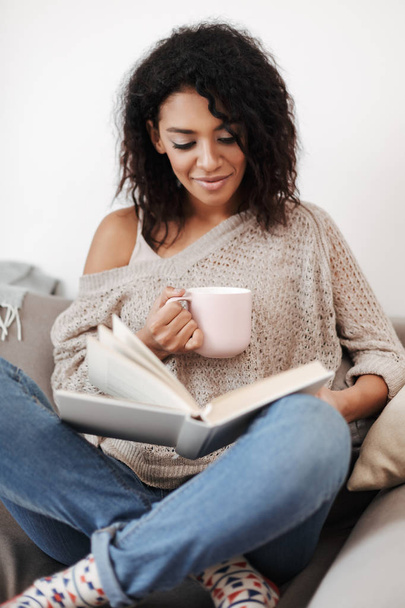 Portrait of young lady with dark curly hair sitting on sofa and reading book with cup in hand at home isolated - Photo, Image