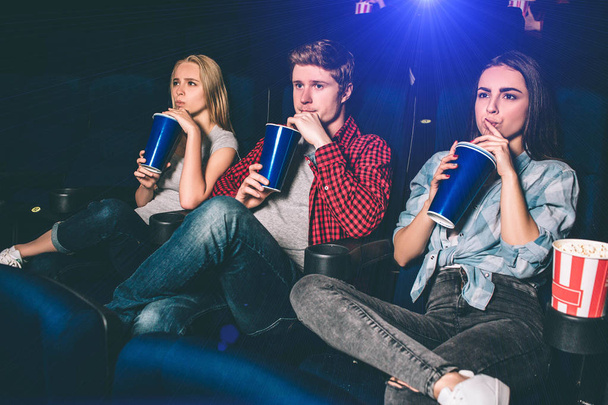 Three friends are sitting together and drinking coke at the same time. They are watching movie. Young people are serious and concentrated on watching. - Fotoğraf, Görsel
