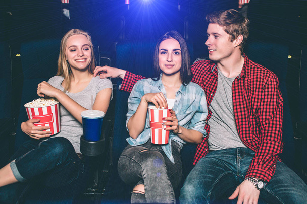 幸せな友人は、映画を見ています。彼らは一緒に座っています。男は、ブロンドの女の子を見ています。彼女はそれについて幸せです。ブルネットの少女の楽しみとポップコーンを食べて. - 写真・画像