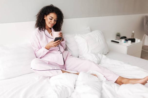 Молодая улыбающаяся леди с темными кудрявыми волосами в сонной одежде сидит в постели с чашкой и сотовым телефоном в руках у себя дома изолированы
 - Фото, изображение