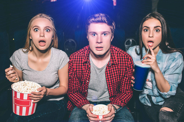 Zdjęcie z zaskoczony i zdumiony Młodzi ludzie patrząc na kamery. Blond dziewczyna trzyma koszyka popcorn a brunetka dziewczyna ma colę w jej ręce. Mężczyzna siedzi między nimi. - Zdjęcie, obraz