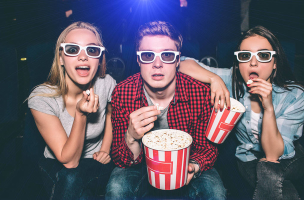 Beautiful people in glasses are looking straight forward. They has opened their mouthes. Each of them has a piece of popcorn in their hands. They look excited. - Photo, image