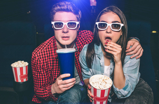 Portrait of two people sitting together in cinema hall and wearing glasses. Girl is amazed and eating popcorn while her partner is drinking cola. - Foto, Bild