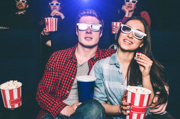 Twee jonge mensen zitten dicht bij elkaar. Ze hebben speciale bril op hun gezichten om film te bekijken. Meisje houdt mand met popcorn en een stuk van het in haar handen. Man heeft een kopje van cokes. - Foto, afbeelding