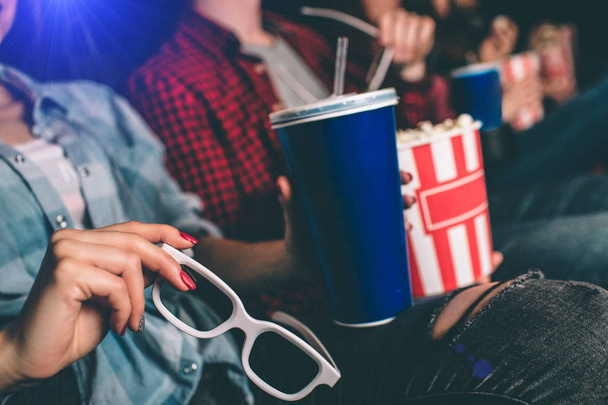 Close up of girls hands keeping glasses and a blue cup of coke. Her partner in red shirt is holding a basket of popcorn. - Foto, Imagem
