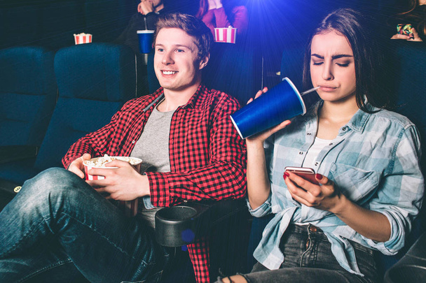 Distracted girl is watching on the phone. She is bored with watching movie. Also she is drinking coke from big and blue cup. Guy is happy and watching movie. - Foto, imagen