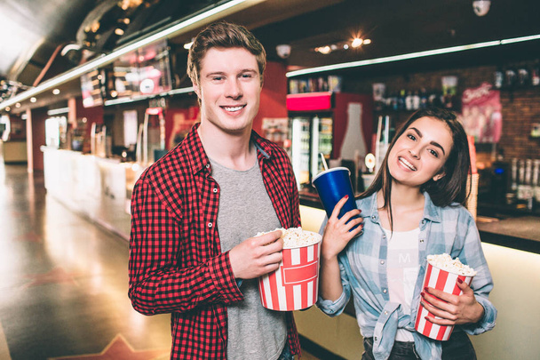 Handsome guy and beautiful girl are posing. They are looking straight and smiling. Guy has a basket of popcorn while girl is holding a cup of coke and basket of popcorn. - Фото, изображение