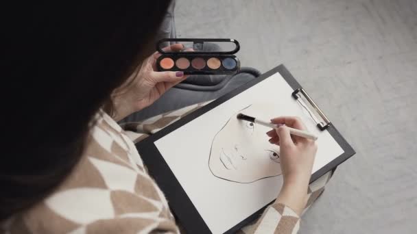 Top view. Young woman makeup artist in a bright studio draws a face chart. The make-up artist draws a schematic image of a face on watercolor paper and using shadow palette and brush cosmeticks - Metraje, vídeo