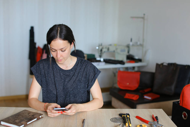 Δημιουργικό craftswoman κάθεται στο ατελιέ και χρησιμοποιώντας το smartphone, χειροποίητα δερμάτινα είδη. - Φωτογραφία, εικόνα