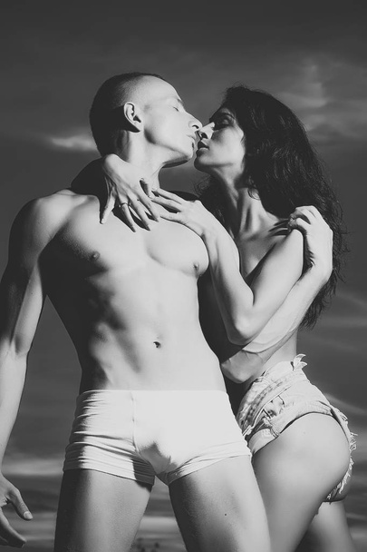 Kissing couple. Sexy young undressed couple of muscular boy and brunette girl with straight beautiful body standing embracing close to each other in sun light on blue sky with white clouds background - Zdjęcie, obraz