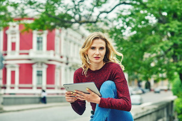 Young woman with tablet on city street . Stylish young woman in jeans and Burgundy sweater uses modern technology - 写真・画像