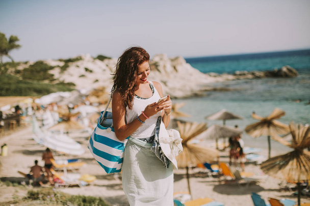 Jolie jeune femme avec un sac utilisant un téléphone portable sur la plage à la journée ensoleillée d'été
 - Photo, image