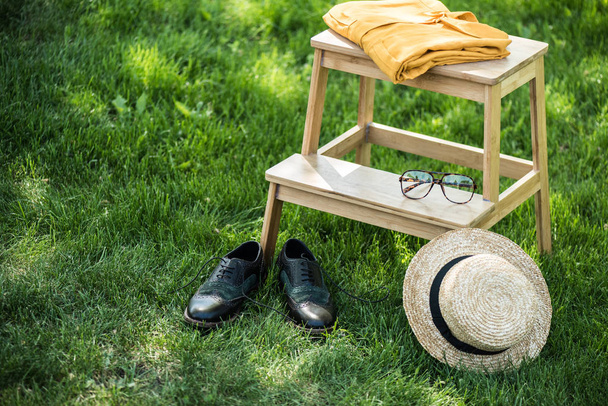 close up view of arrangement of eyeglasses, black shoes, shirt and hat on wooden stairs on green grass - Photo, Image