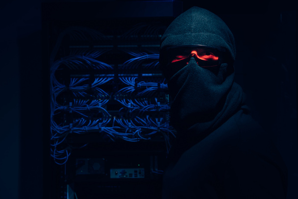 vista oscurecida de hacker en gafas y máscara mirando a la cámara con cables en el fondo
 - Foto, imagen