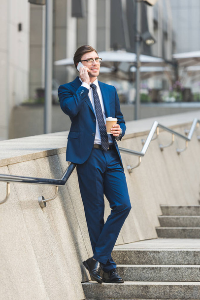 successful young businessman in stylish suit with coffee to go talking by phone on stairs near business building - Photo, Image