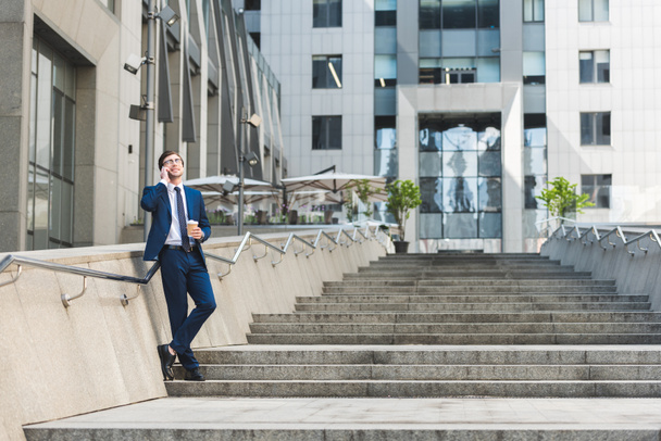 handsome young businessman in stylish suit with coffee to go talking by phone on stairs near business building - Foto, Imagen