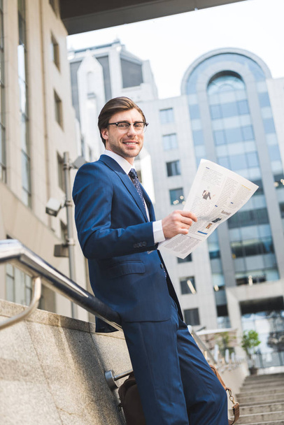 bottom view of smiling young businessman in stylish suit with newspaper in business district - Foto, Imagen