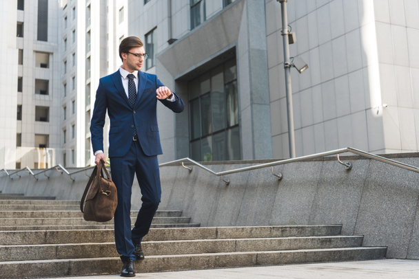 attraktiver junger Geschäftsmann im schicken Anzug mit Aktentasche, der auf die Uhr im Geschäftsviertel schaut - Foto, Bild