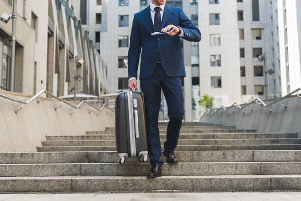 tiro cortado de homem de negócios em terno elegante com bagagem e bilhetes de voo descendo escadas no distrito de negócios
 - Foto, Imagem