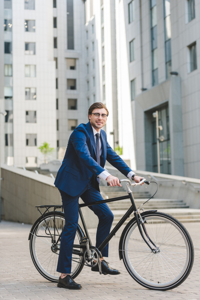 schöner junger Geschäftsmann im schicken Anzug auf dem Fahrrad im Geschäftsviertel - Foto, Bild