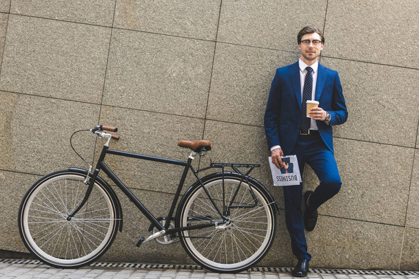 schöner junger Geschäftsmann im schicken Anzug mit Zeitung und Fahrrad an der Wand gelehnt - Foto, Bild